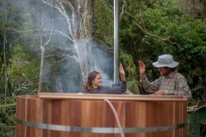Two people high fiving behind a wood fired hot tub in the outdoor wilderness with smoke rising from the wood stove in the background