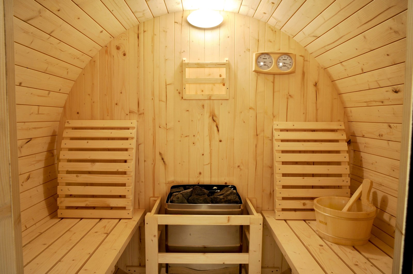 Interior of a Finnish pine sauna with backrest, water bucket, hygrometer, and electric heater with sauna stones