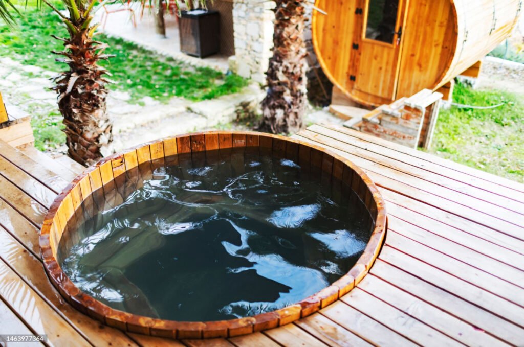 A red cedar hot tub filled with water with trees and a sauna house in the background