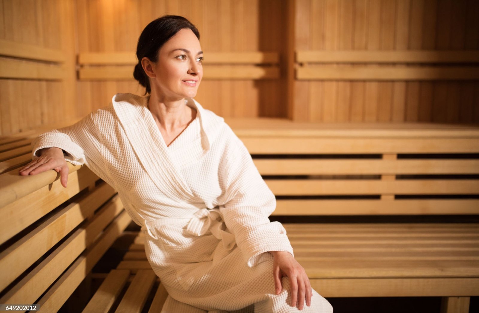 A woman in a white bathrobe sitting inside a sauna, bathed in soft, warm light for a cozy and relaxing atmosphere.