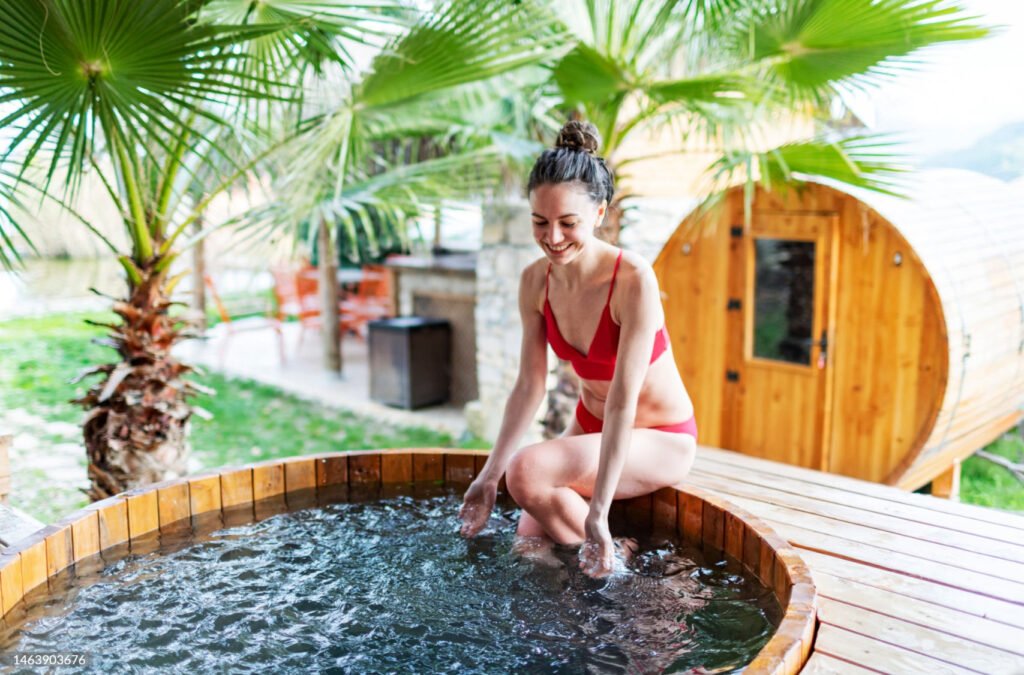 3 Cedar Lover Woman sitting on the edge of a hot tub splashing water with a cedar sauna and elm trees in the background